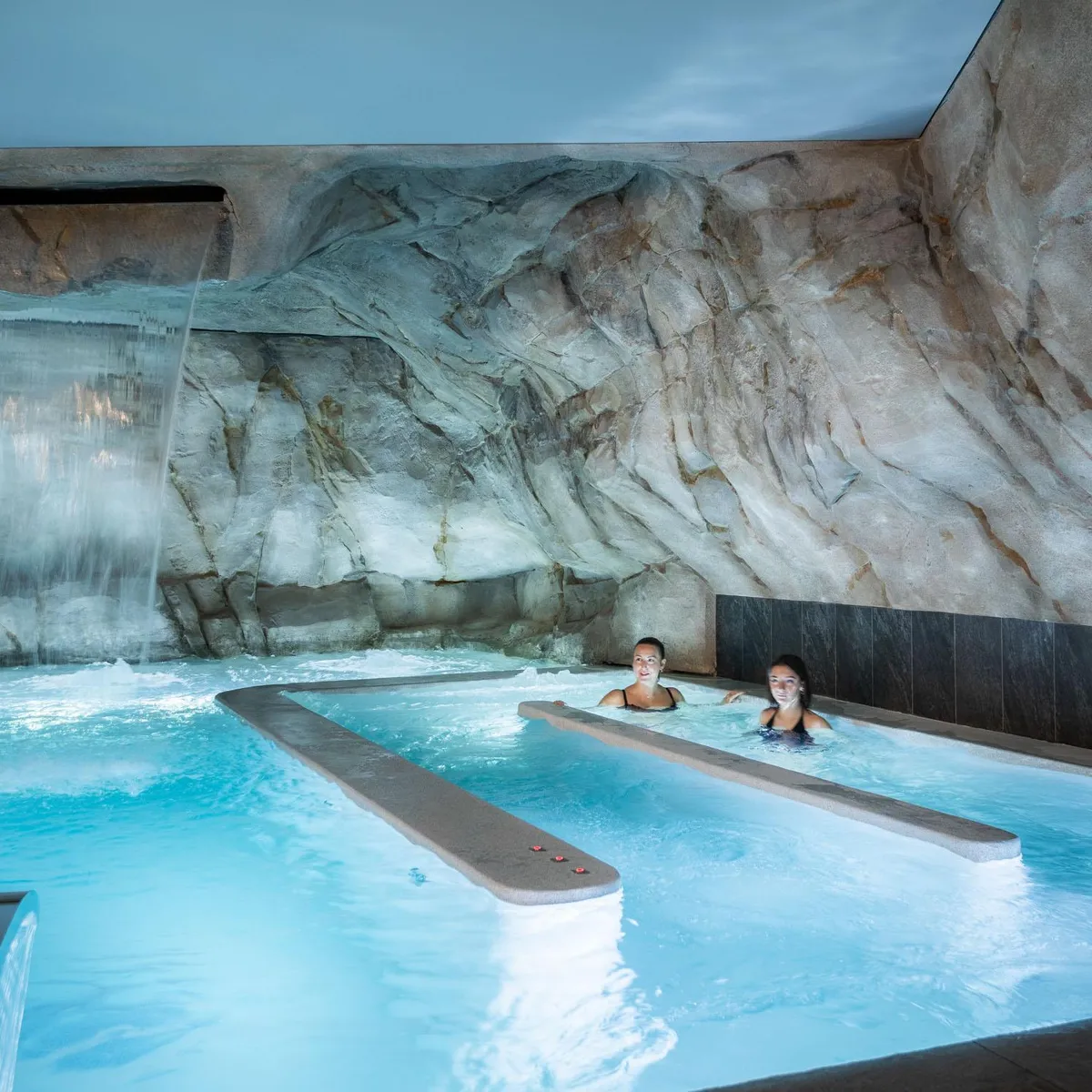 Deux personnes nagent dans une piscine intérieure avec une cascade et des rochers artificiels en arrière-plan