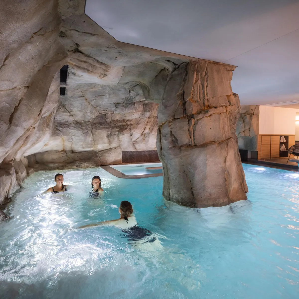 Trois personnes nagent dans une piscine intérieure entourée de parois rocheuses artificielles, avec des chaises longues et des lumières en arrière-plan
