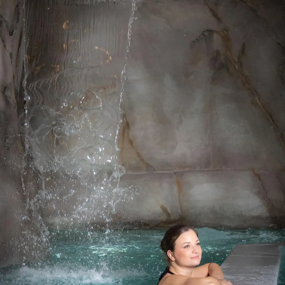 Une femme est allongée dans une piscine avec de l'eau qui coule en cascade sur la paroi rocheuse derrière elle
