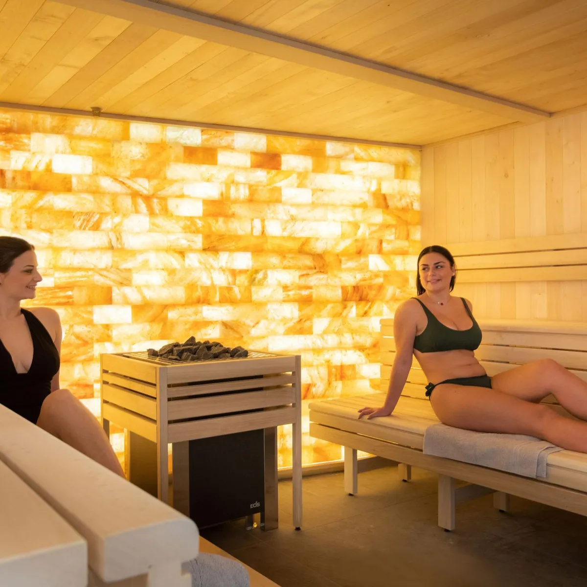 Deux femmes assises sur des bancs en bois dans un sauna éclairé par une paroi lumineuse aux motifs dorés