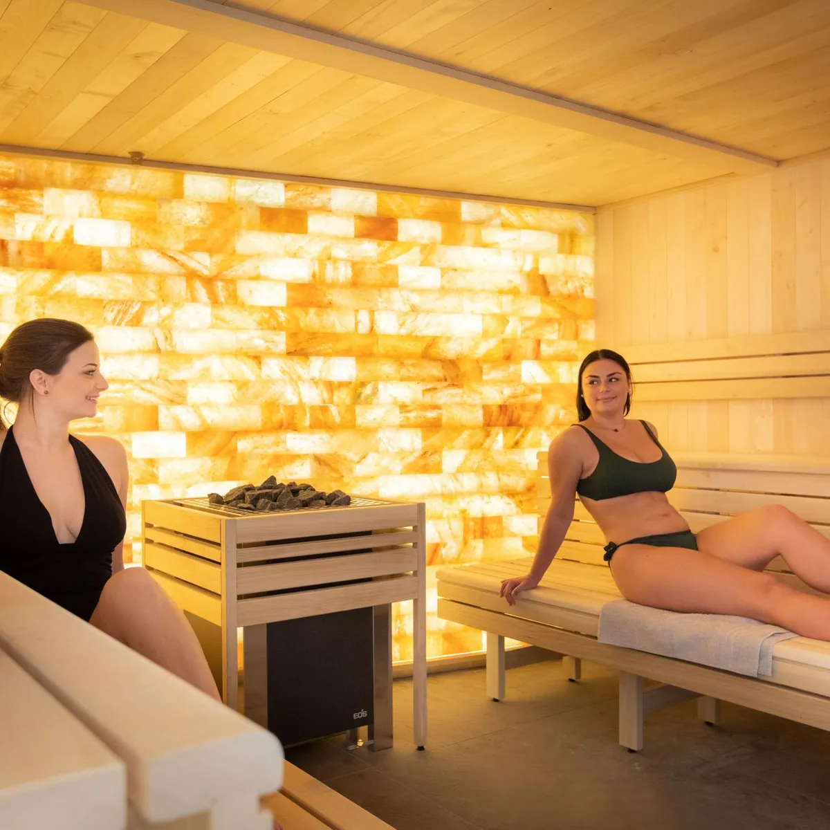 Deux femmes assises dans un sauna en bois avec des pierres chauffantes au centre et un mur lumineux en arrière-plan
