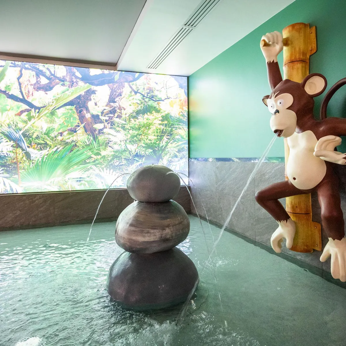 Une sculpture de singe en bois projette de l'eau dans un bassin avec des rochers et une peinture murale représentant une jungle