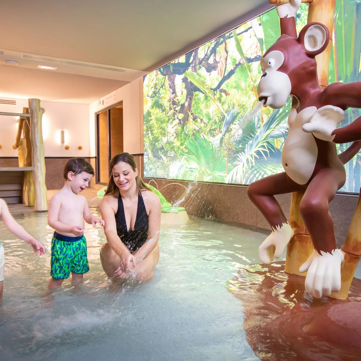 Une femme et deux enfants jouent dans une piscine intérieure avec un décor de jungle et une sculpture d'animal