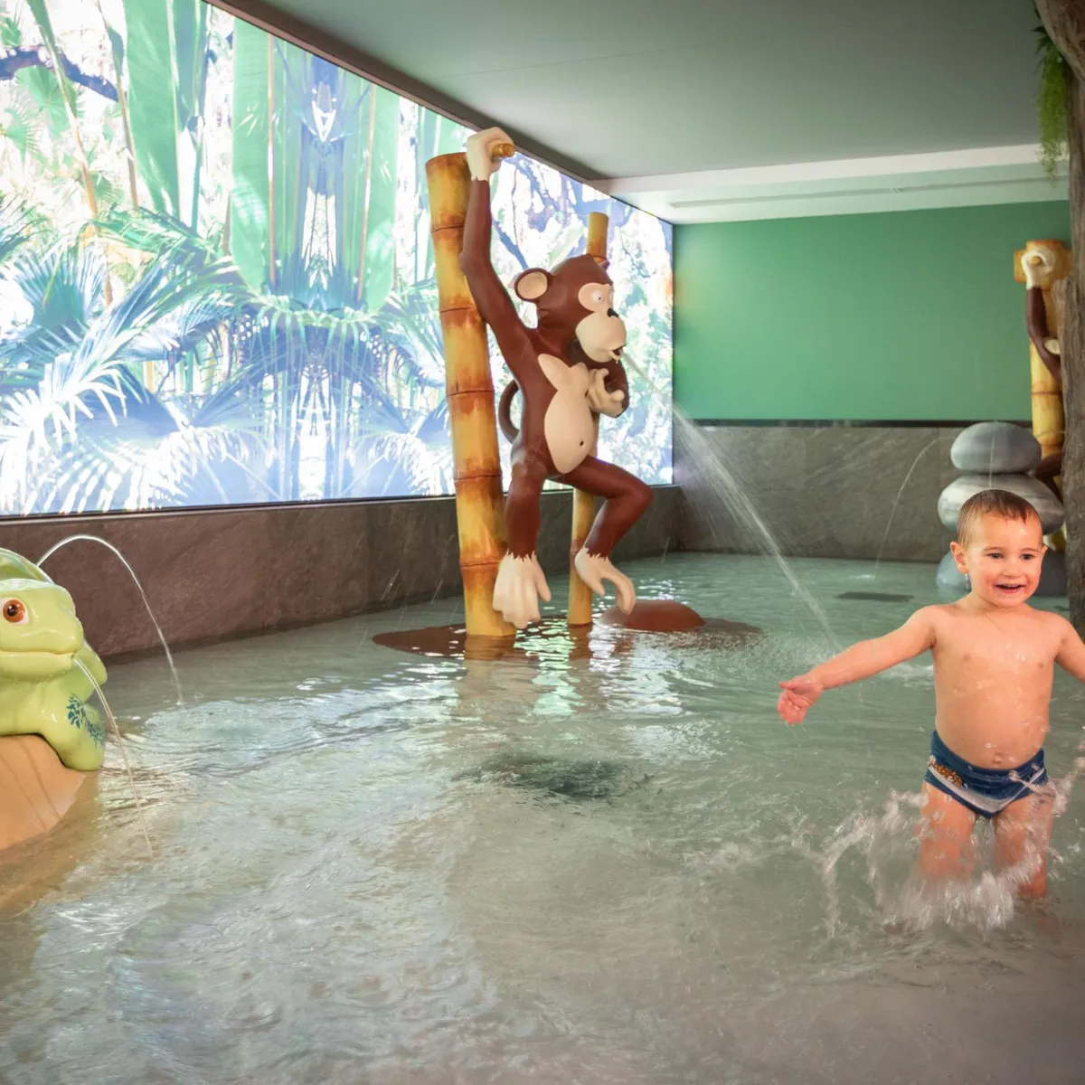 Enfant jouant dans une piscine pour enfants avec des sculptures d'animaux et des jets d'eau, décorée de motifs de jungle