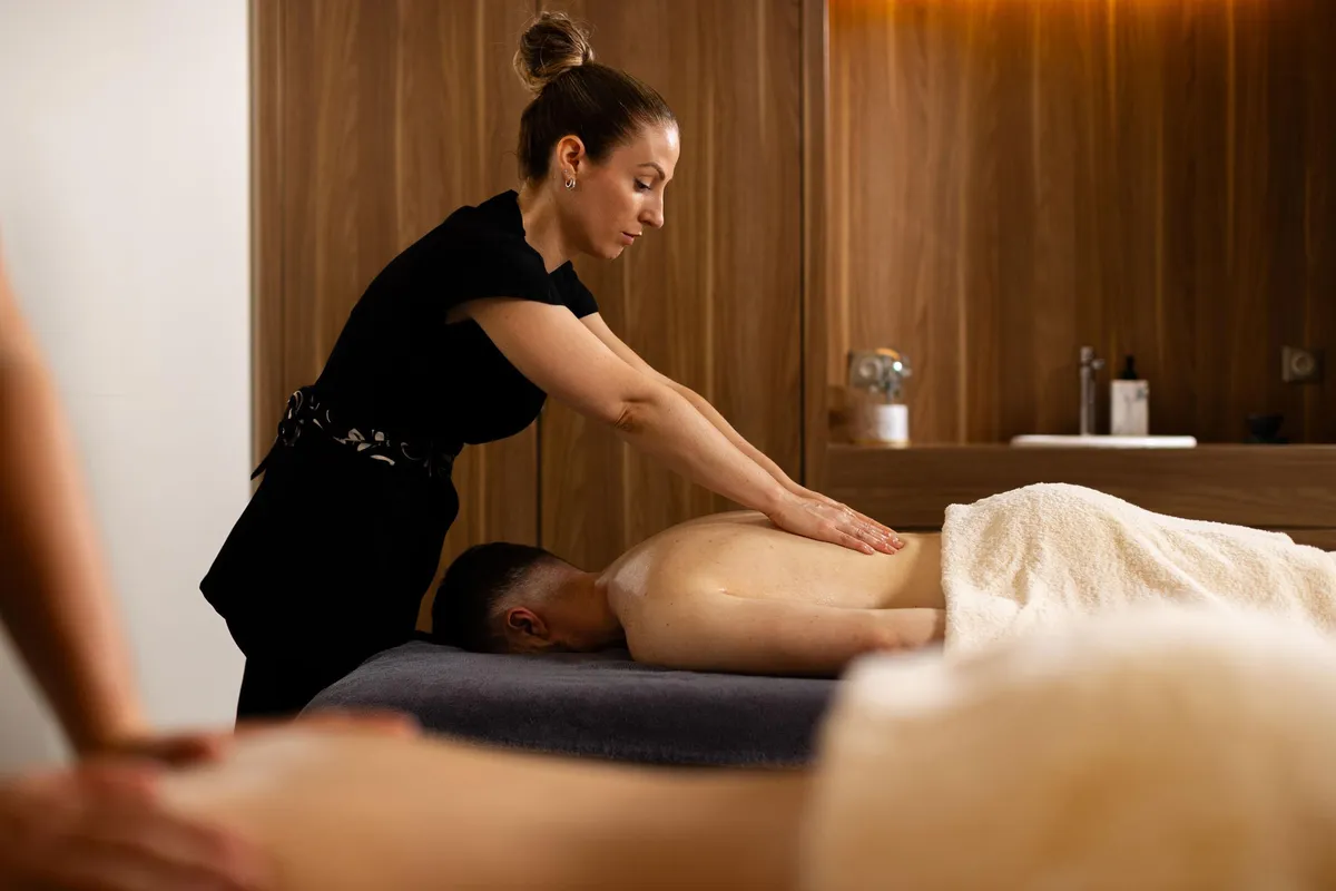Eine Frau in schwarzer Kleidung massiert eine Person, die auf dem Bauch auf einer mit einem Handtuch bedeckten Massageliege liegt.