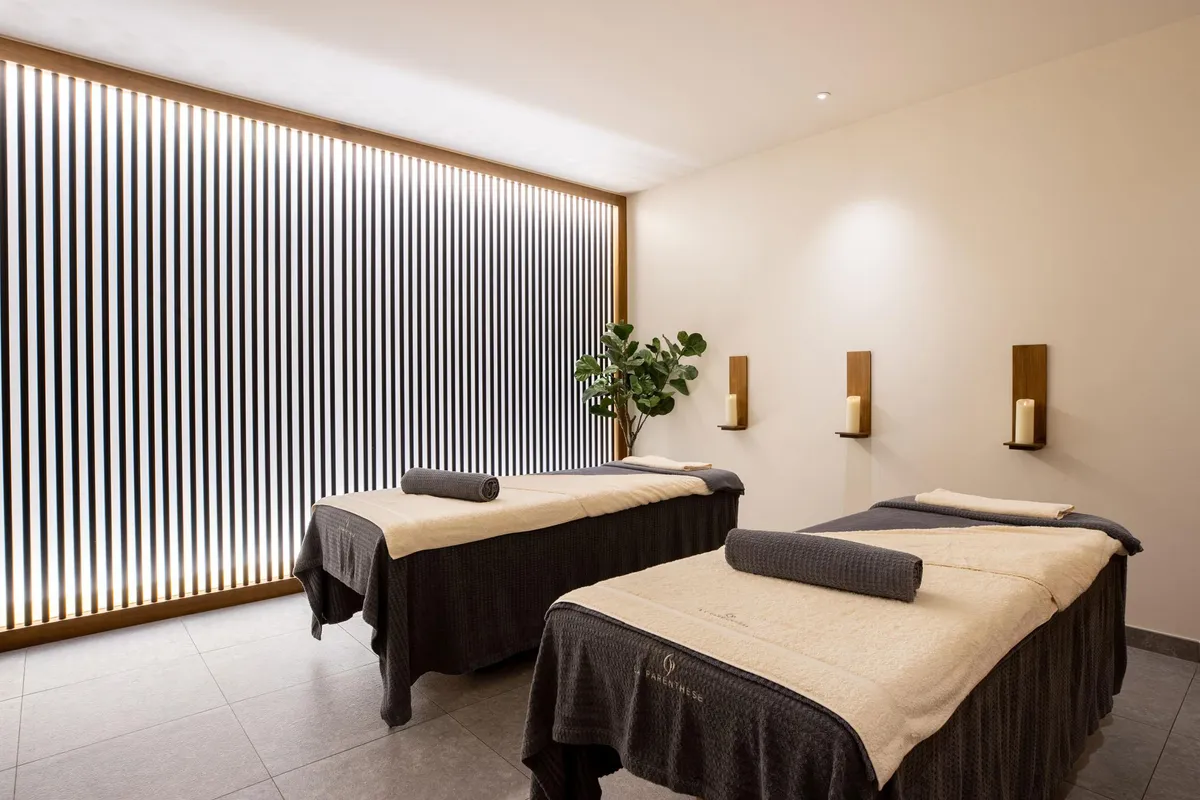 Deux tables de massage avec serviettes, éclairées par des lampes murales, dans une pièce aux murs beiges et à la fenêtre à lamelles