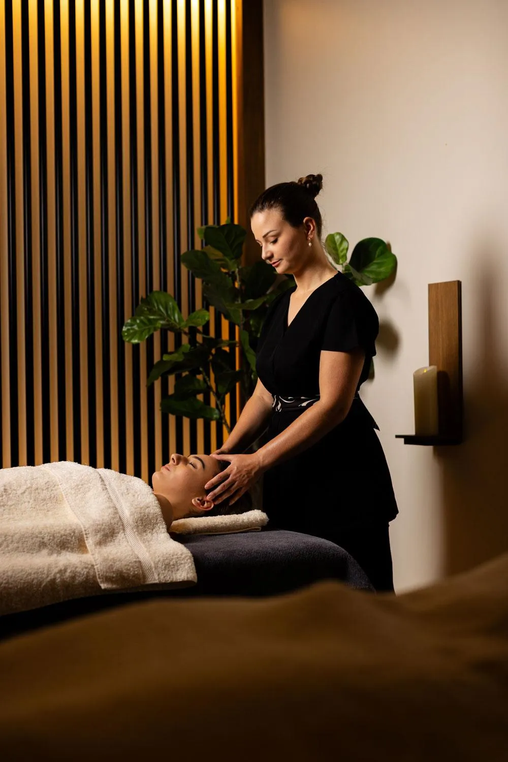 Eine Frau in schwarzer Kleidung führt eine Massage an einer Person durch, die auf einer mit Handtüchern bedeckten Massageliege liegt.
