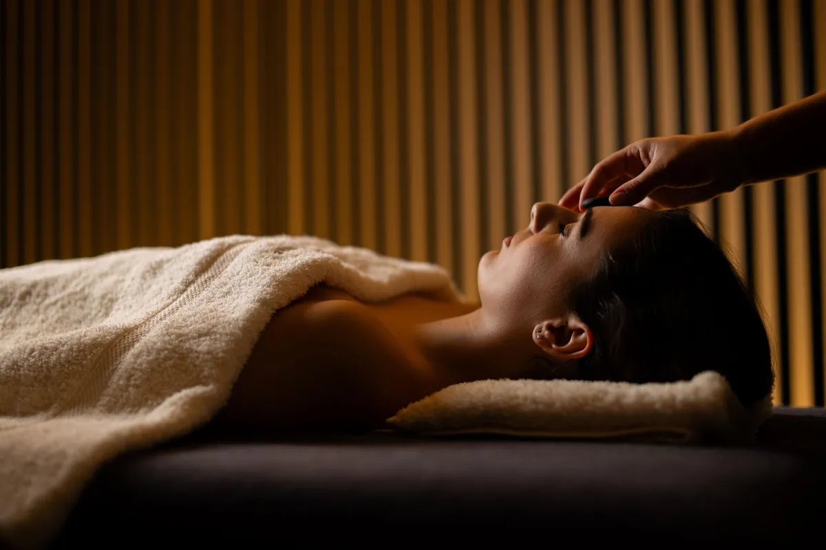 A person lying on a towel receives a facial massage in a room with striped walls.
