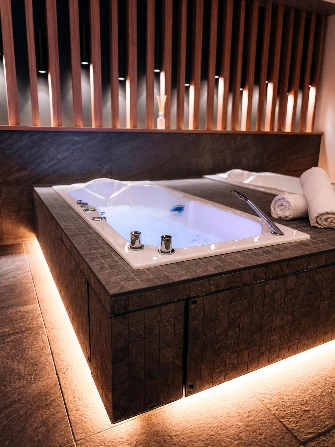 Whirlpool mit Wasser gefüllt, mit LED-Lichtern unter dem Rand, umgeben von dunklem Fliesenboden und weißen Handtüchern