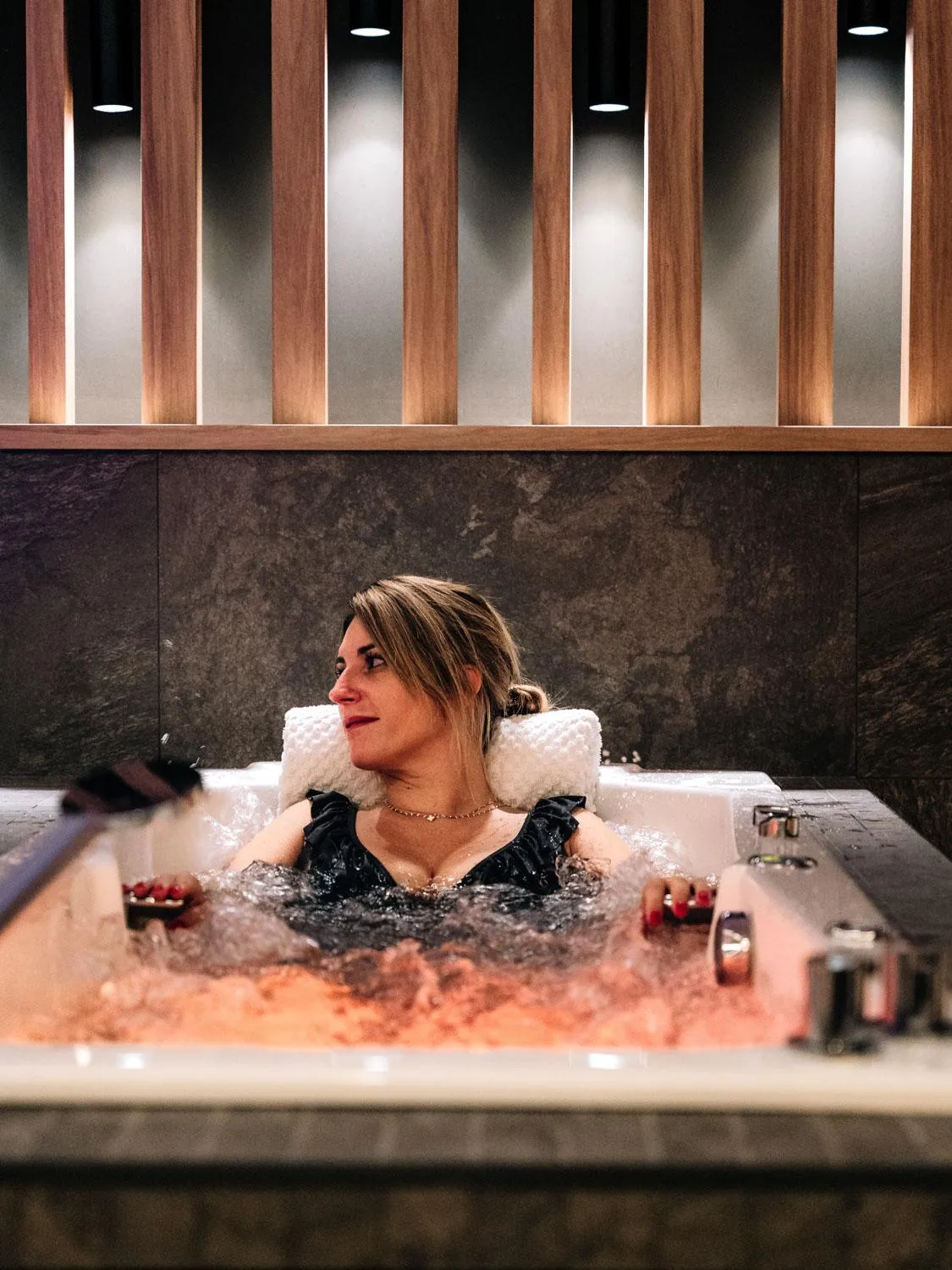 Eine Frau liegt in einem Whirlpool mit einem Fell-Nackenstützkissen, umgeben von Blasen, in einem Innenraum mit Holzsäulen.