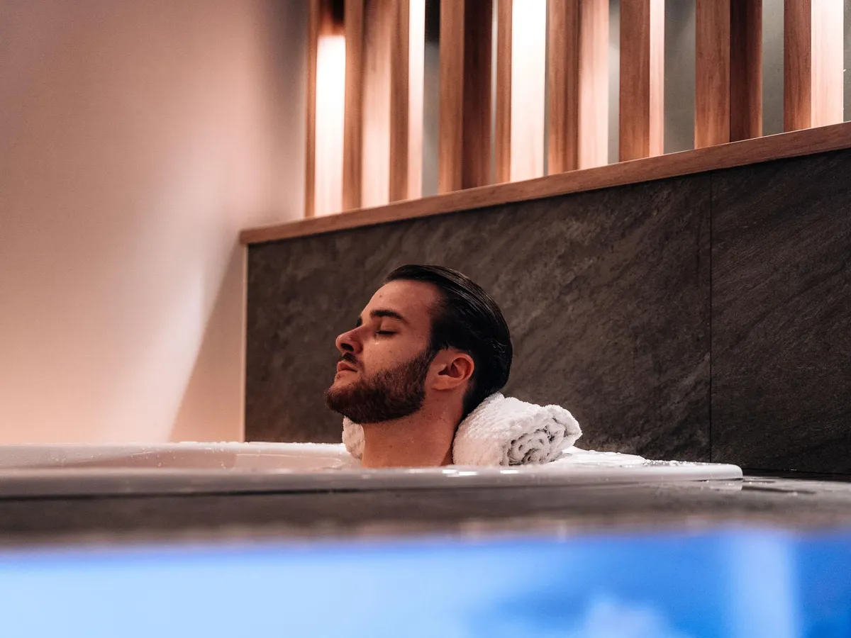 Ein bärtiger Mann liegt in einer Badewanne mit einem weißen Handtuch über den Schultern und geschlossenen Augen.