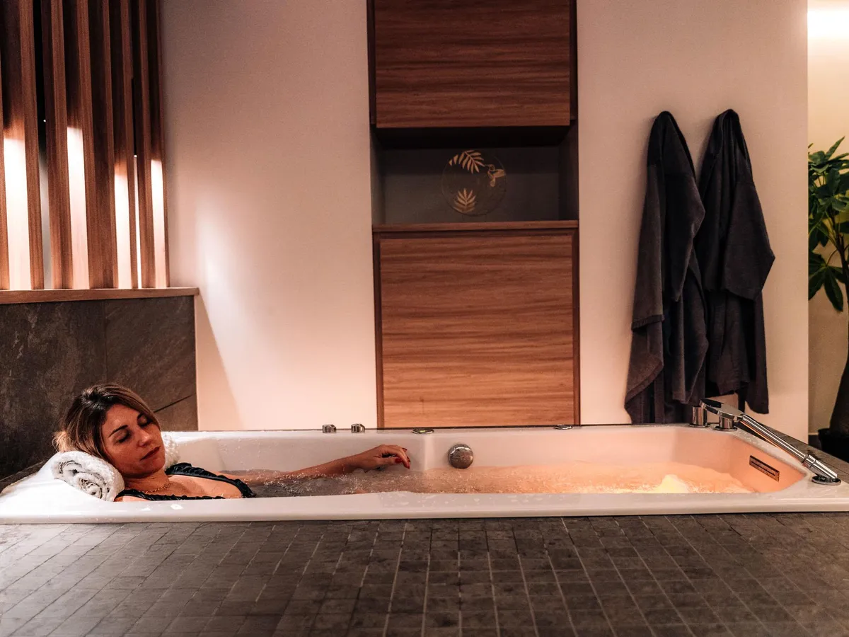 Eine Person liegt in einer mit Wasser gefüllten Badewanne, mit Handtüchern am Rand und Bademänteln in der Nähe aufgehängt.