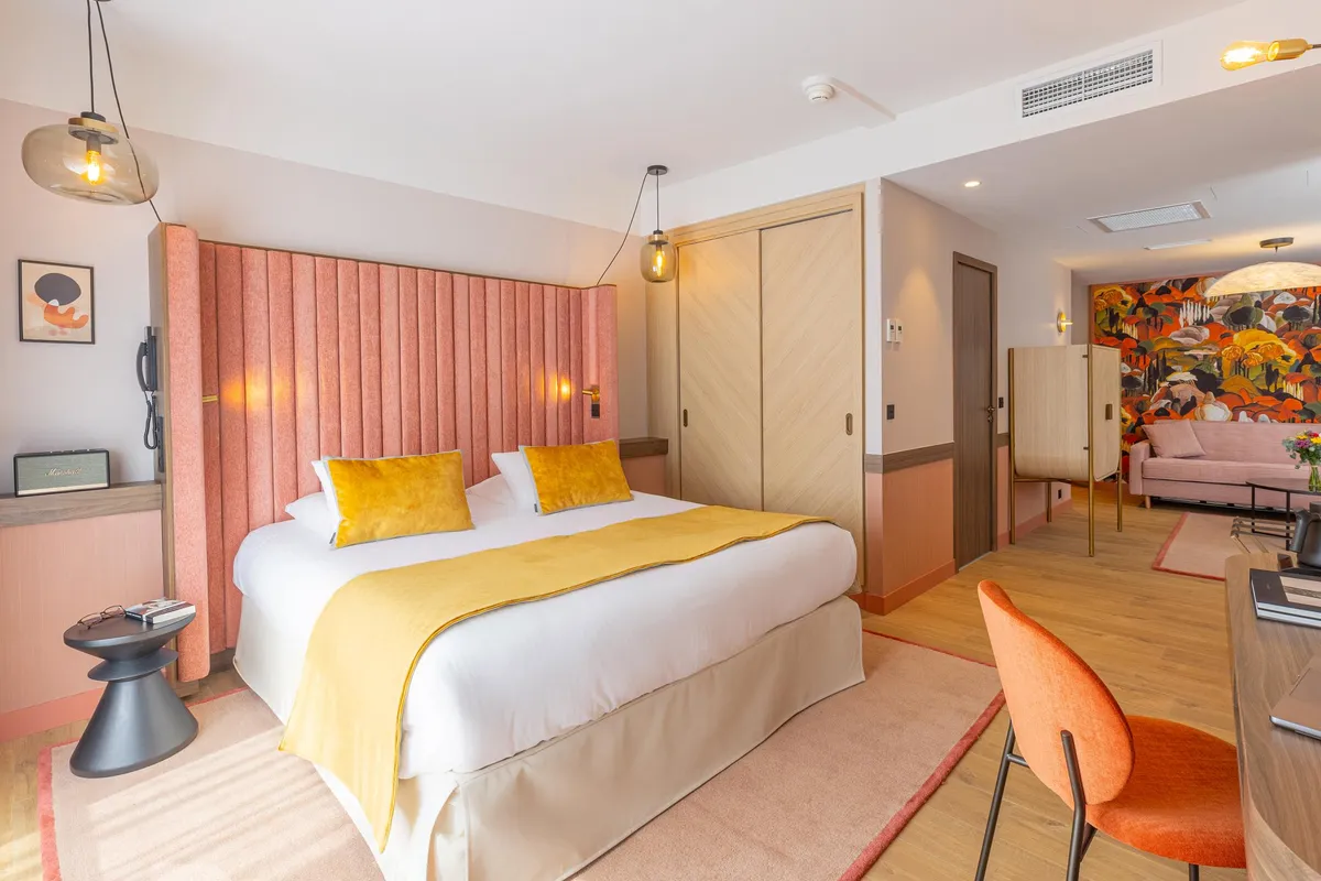 Hotelzimmer mit einem Doppelbett mit weißen Laken und einer gelben Decke, einem rosa Kopfteil, einem Nachttisch mit Gegenständen darauf, einer Hängelampe, einem orangen Sessel und einer offenen Tür zu einem Wohnzimmer mit einem Sofa und einem Wandbild