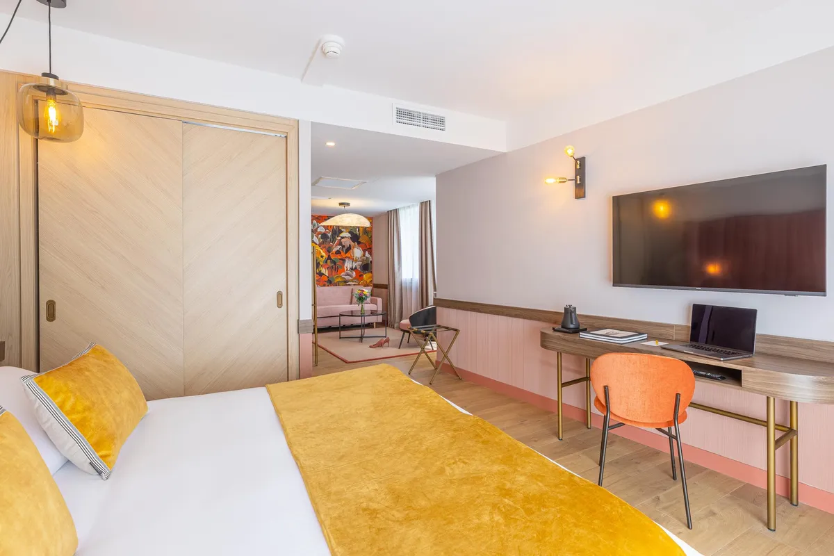 Modernes Hotelzimmer mit Bett, Schreibtisch, Fernseher und sichtbarer Loungefläche im Hintergrund