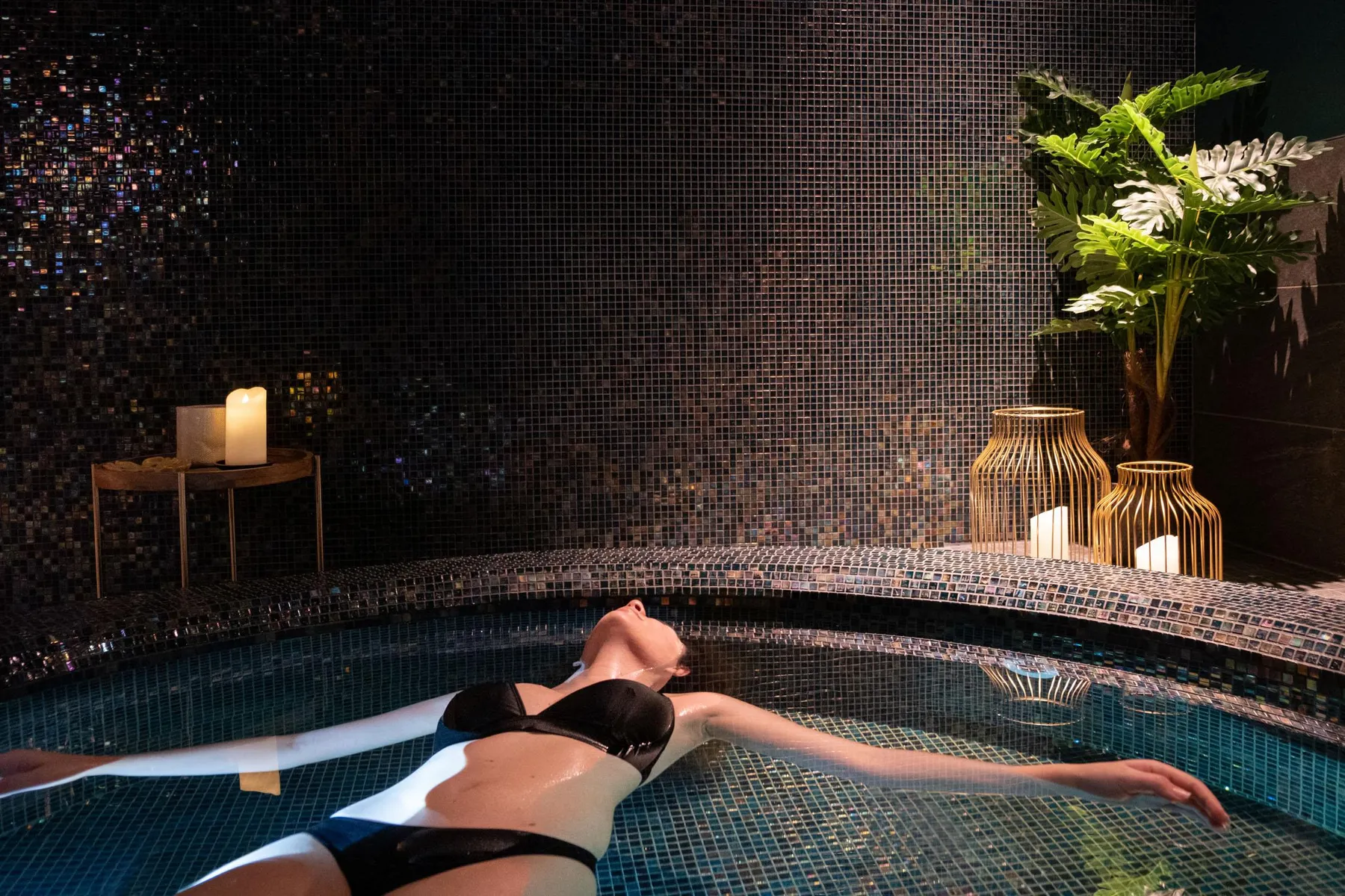 Une personne en maillot de bain noir flotte sur le dos dans une piscine intérieure avec une mosaïque murale et des décorations lumineuses