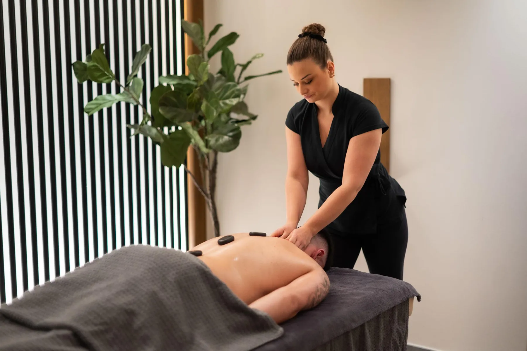 Une femme en tenue noire pratique un massage sur le dos d'une personne allongée sur une table recouverte d'une serviette, avec une plante verte à proximité