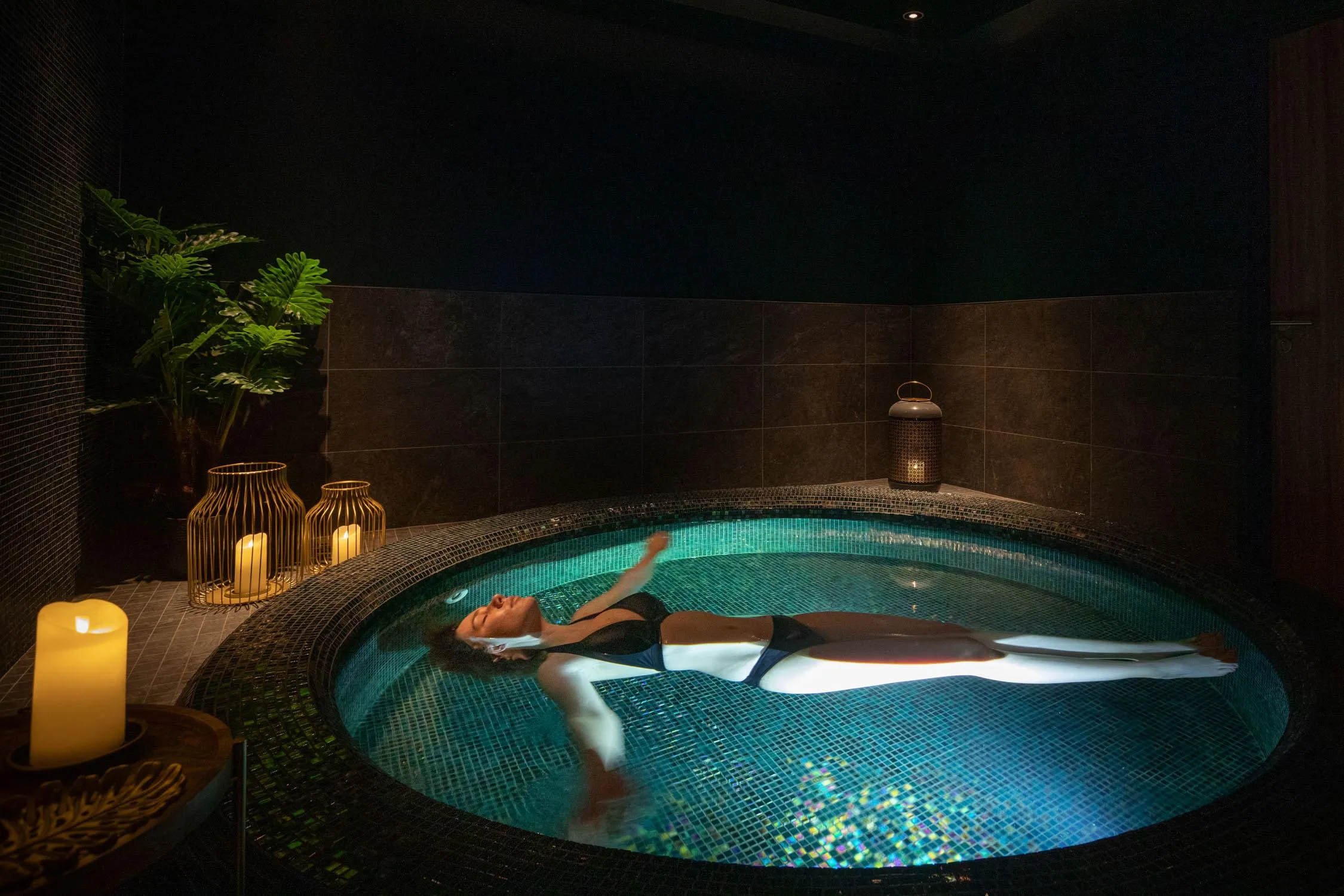 A person is lying in a circular hot tub surrounded by candles and green plants.