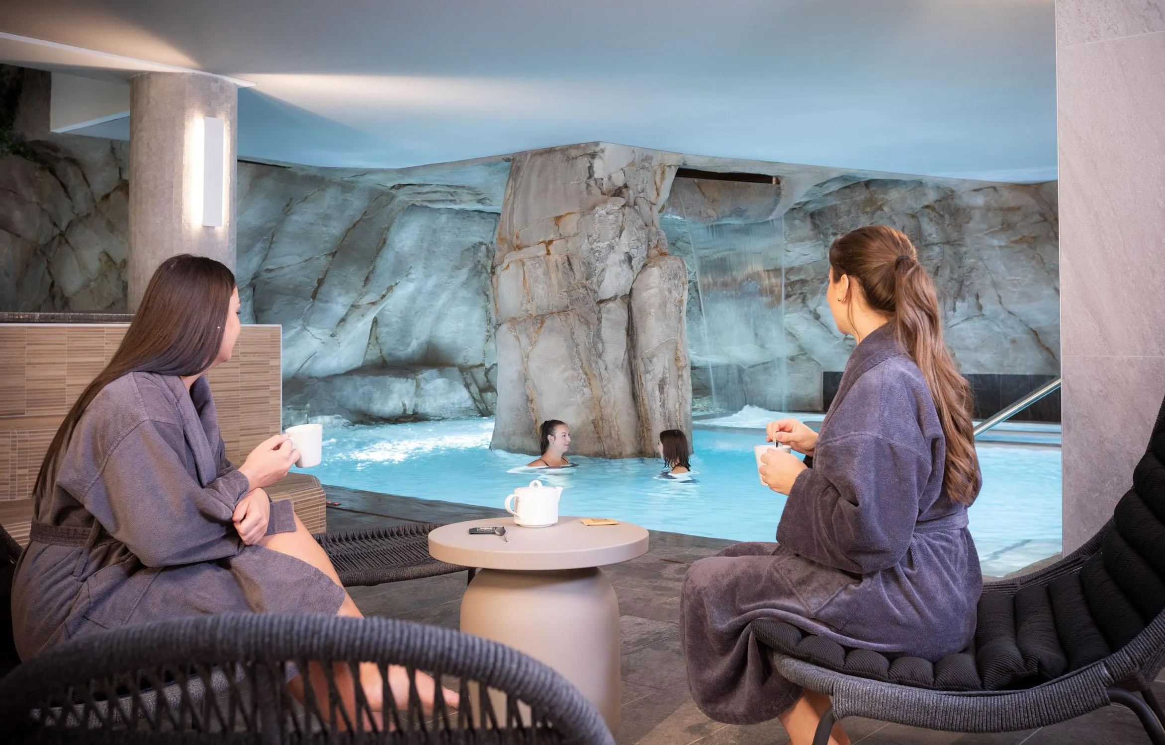 Deux personnes assises en robe de bain discutent près d'une piscine intérieure avec cascade et rochers artificiels, tenant des tasses, tandis que d'autres personnes nagent