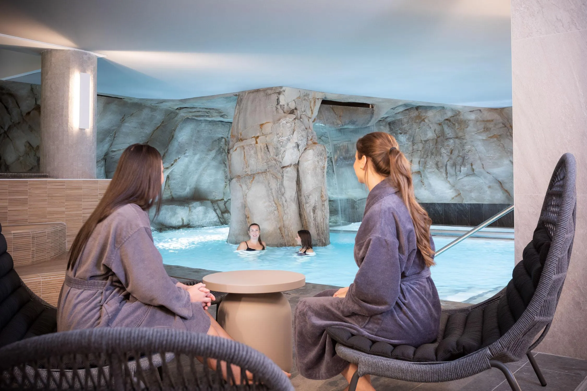 Deux personnes assises en robe de bain observent un bassin d'eau avec des rochers et des cascades, où deux autres personnes sont en train de nager