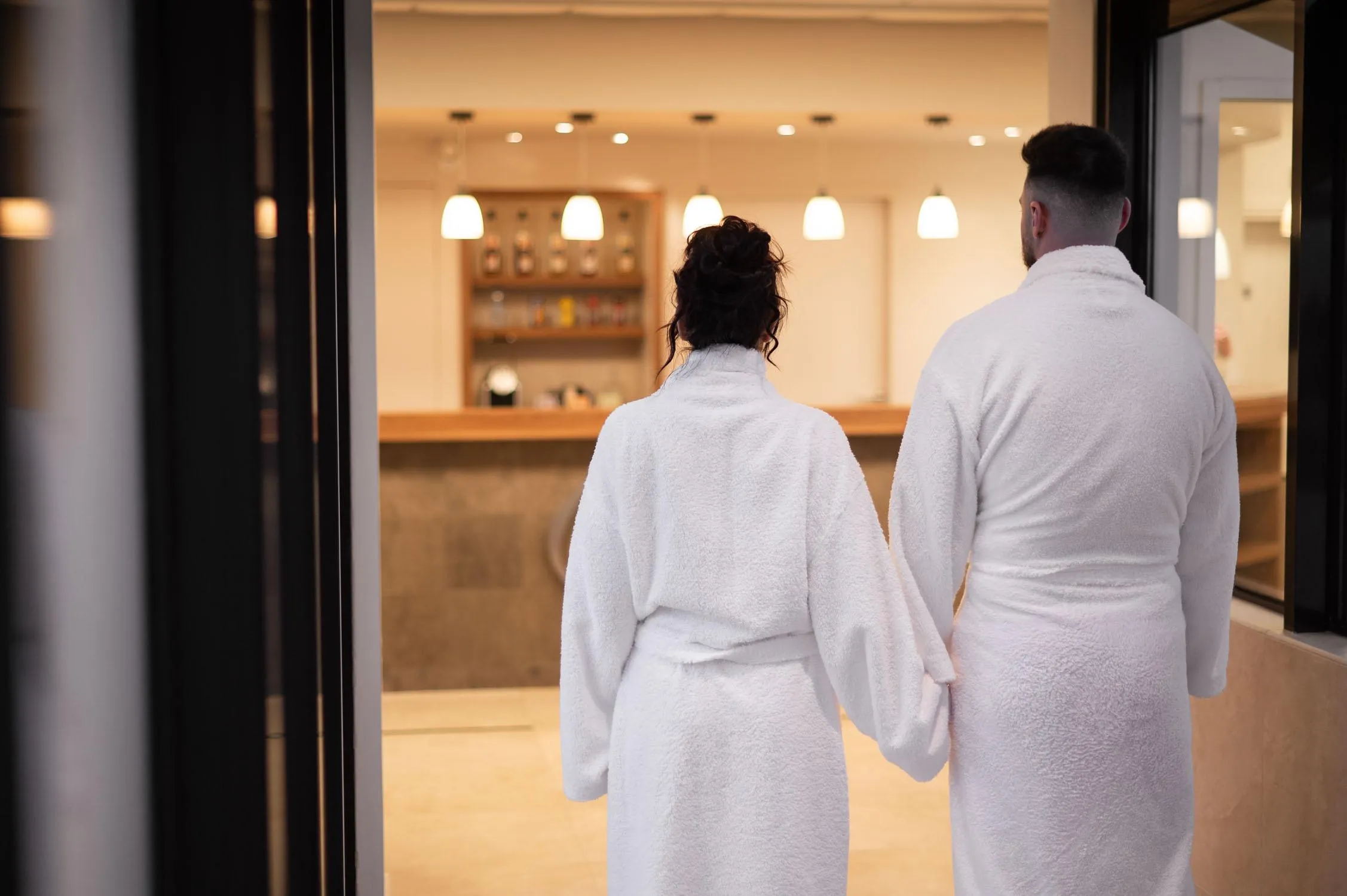 Un couple en peignoirs blancs se tient dos à la caméra devant une vitrine de spa, main dans la main, observant l'intérieur éclairé