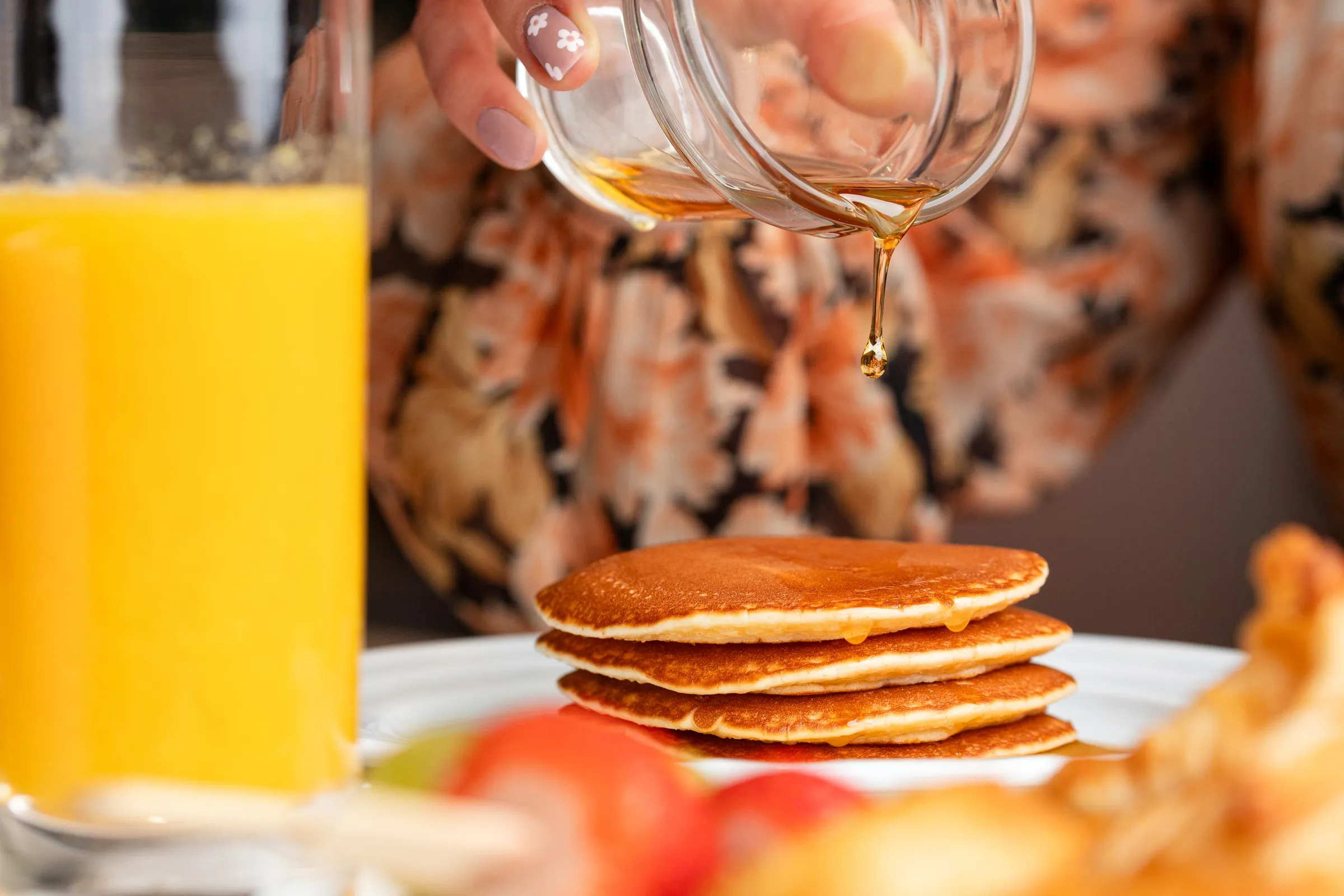 Une main verse du sirop sur une pile de pancakes, avec un verre de jus d'orange à gauche