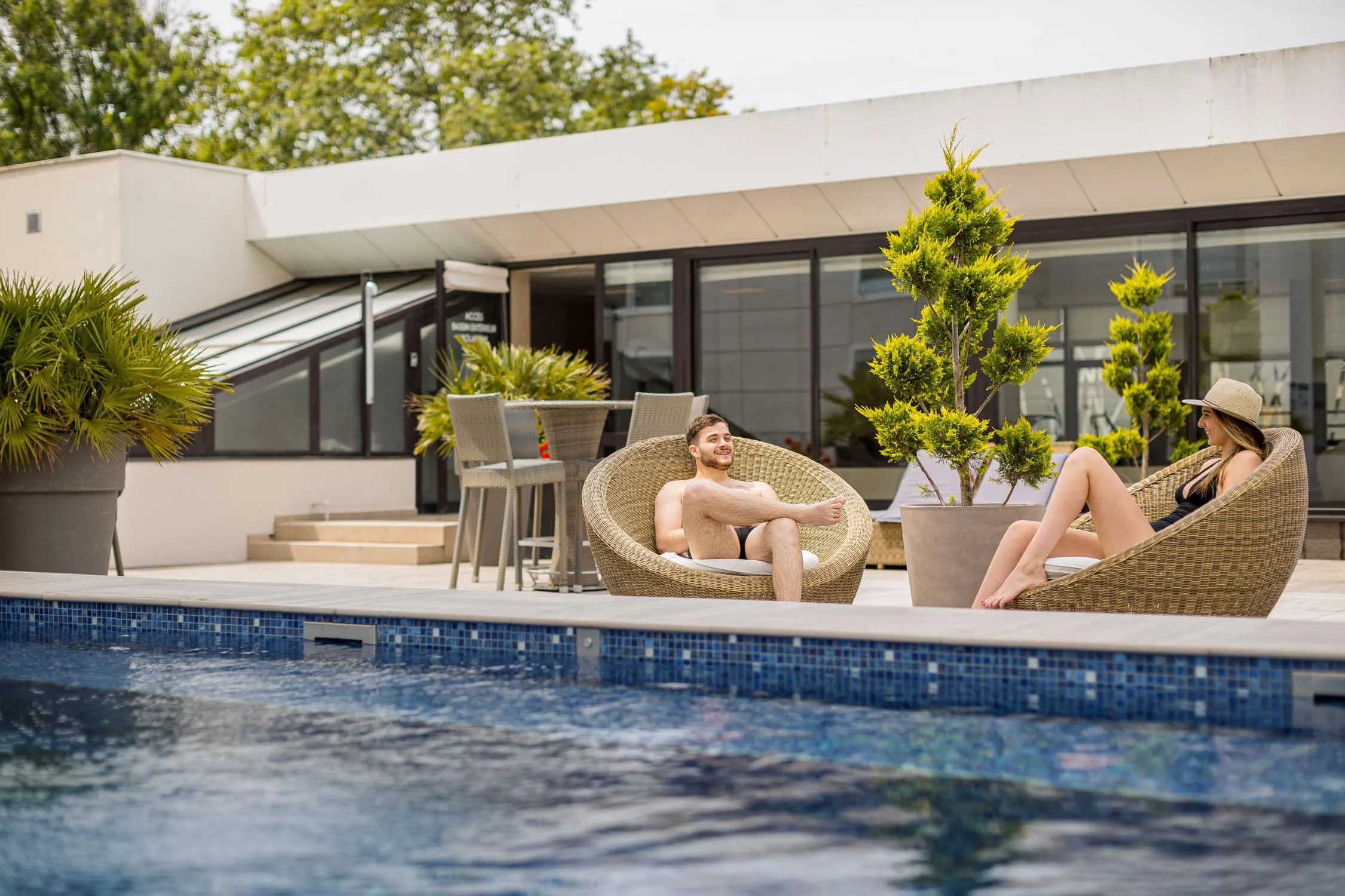 Deux personnes assises dans des fauteuils en osier au bord d'une piscine devant un bâtiment moderne avec des plantes en pot