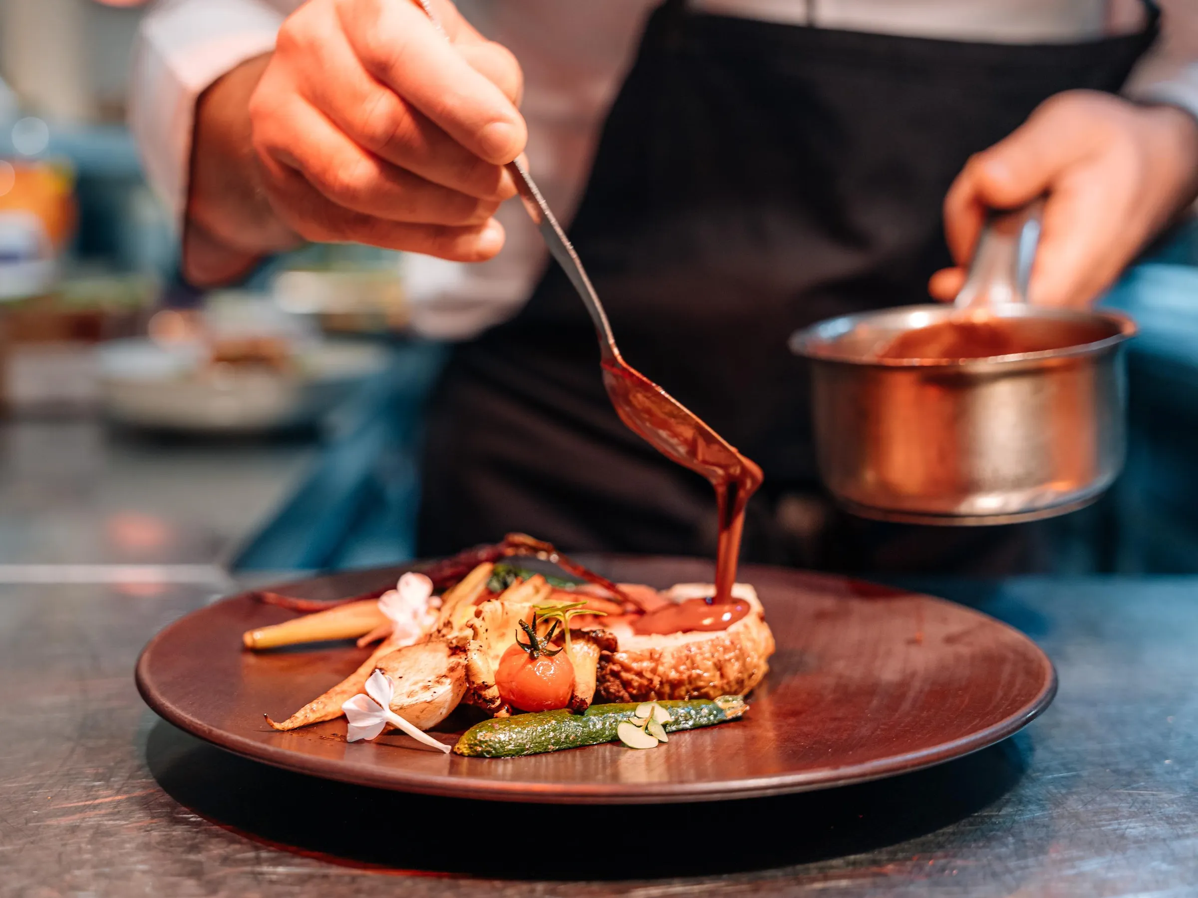 Un chef cuisine verse une sauce sur un plat composé de légumes et de viande grillée