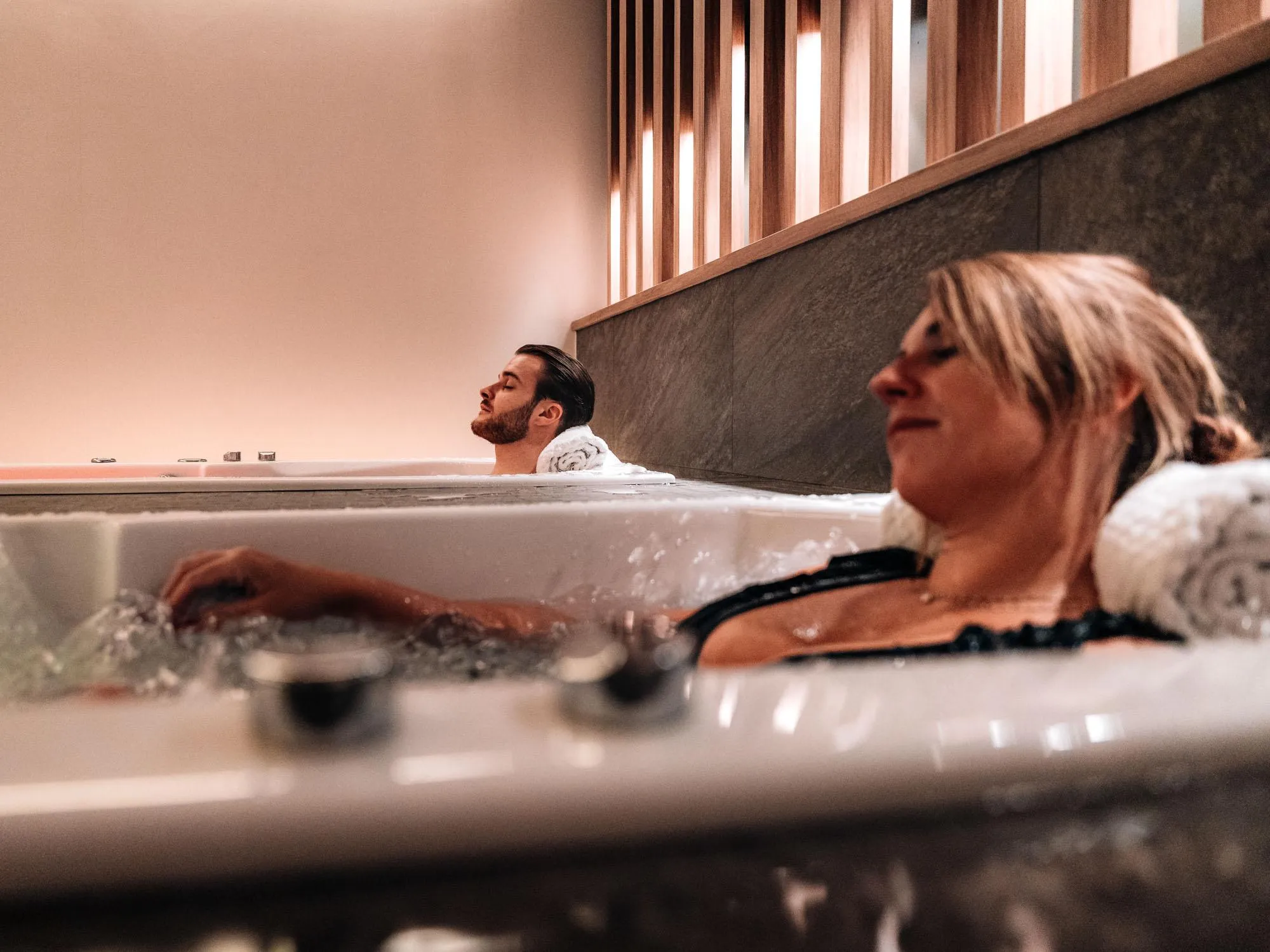 Two people are lying in separate baths, eyes closed, in a space with stone walls and wooden windows.