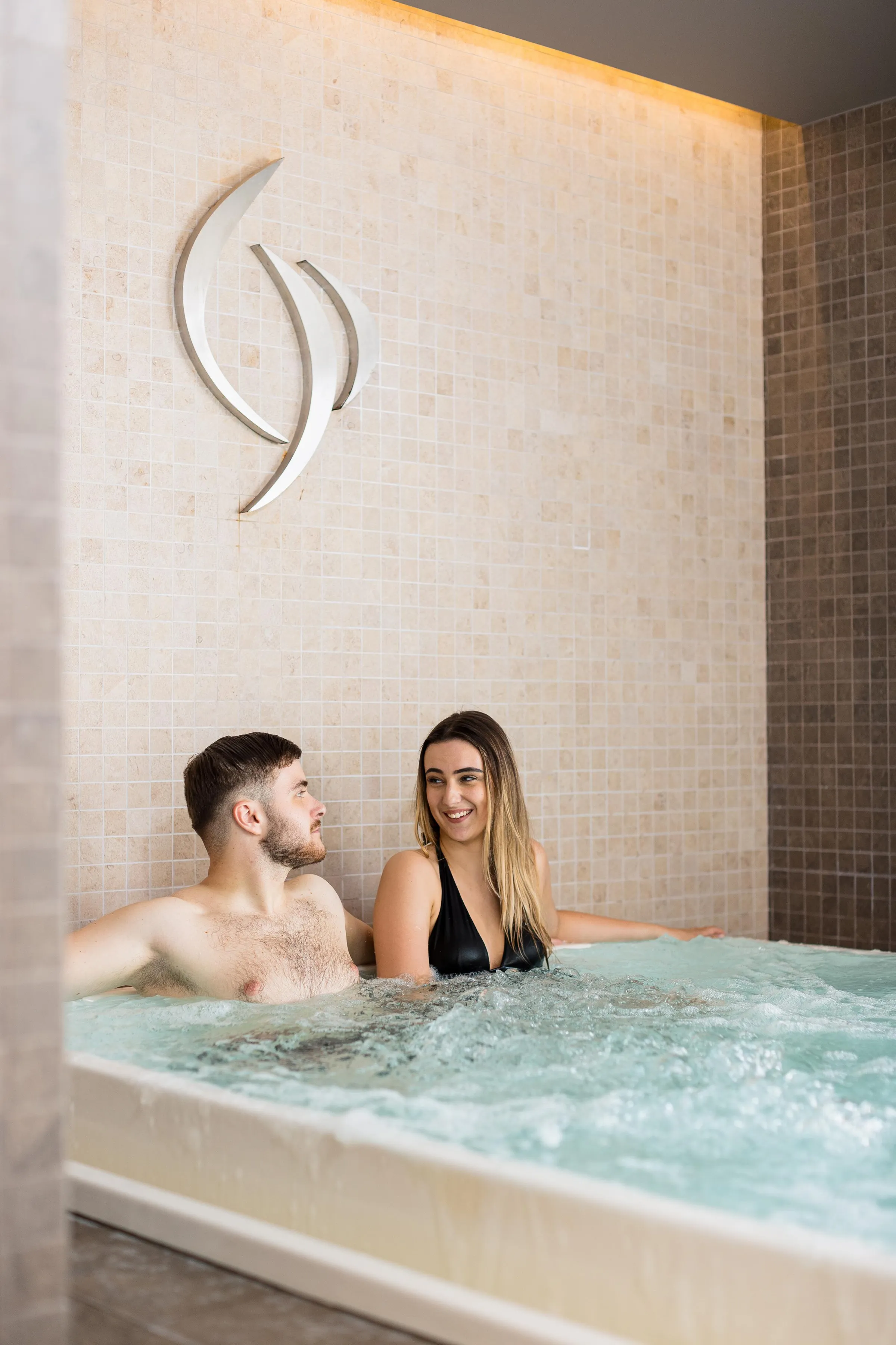 Un homme et une femme sourient dans un bain à remous avec un symbole de lune au-dessus du mur carrelé