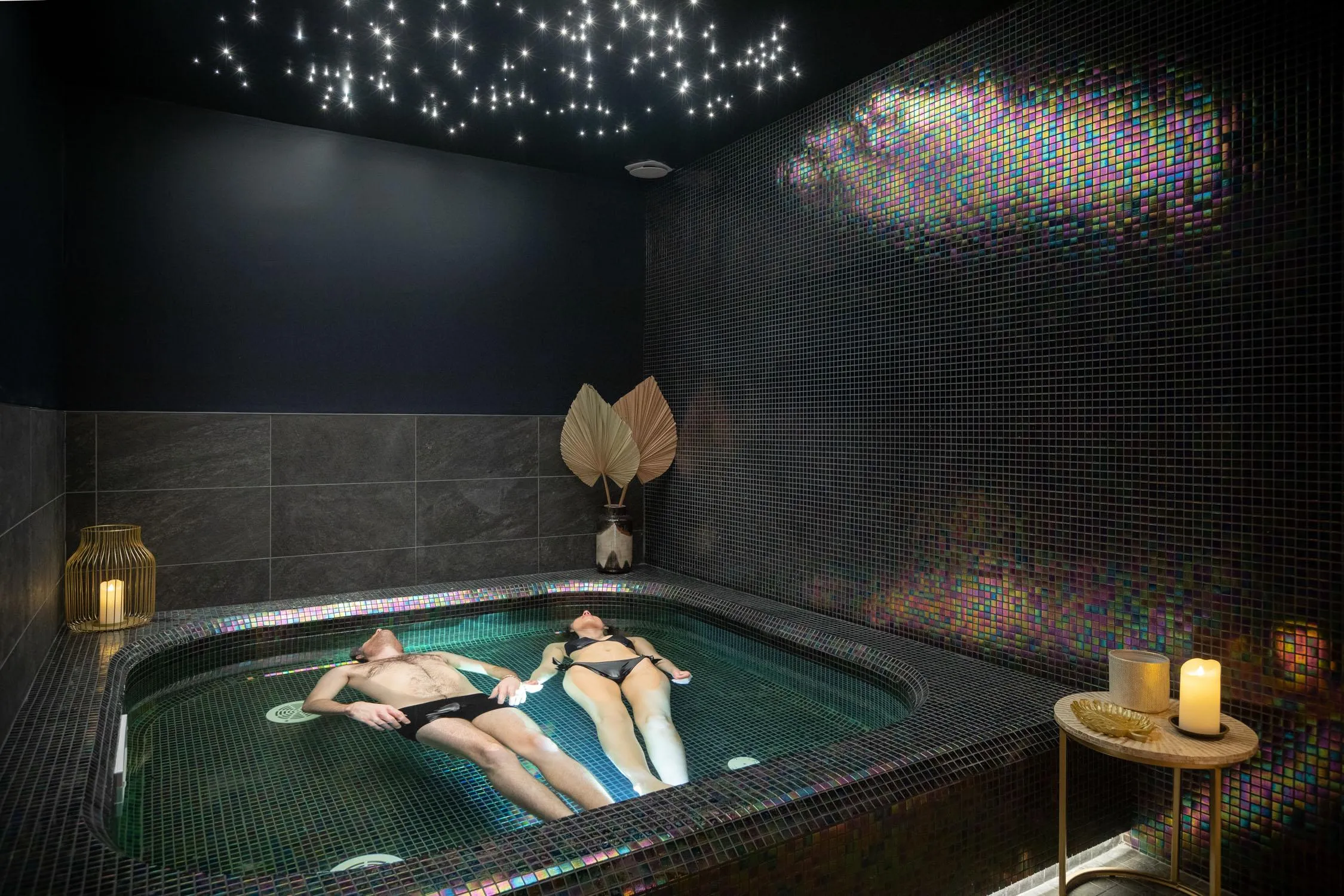 Deux personnes allongées dans un bain à remous, entourées de mosaïques colorées et de bougies, avec un plafond étoilé
